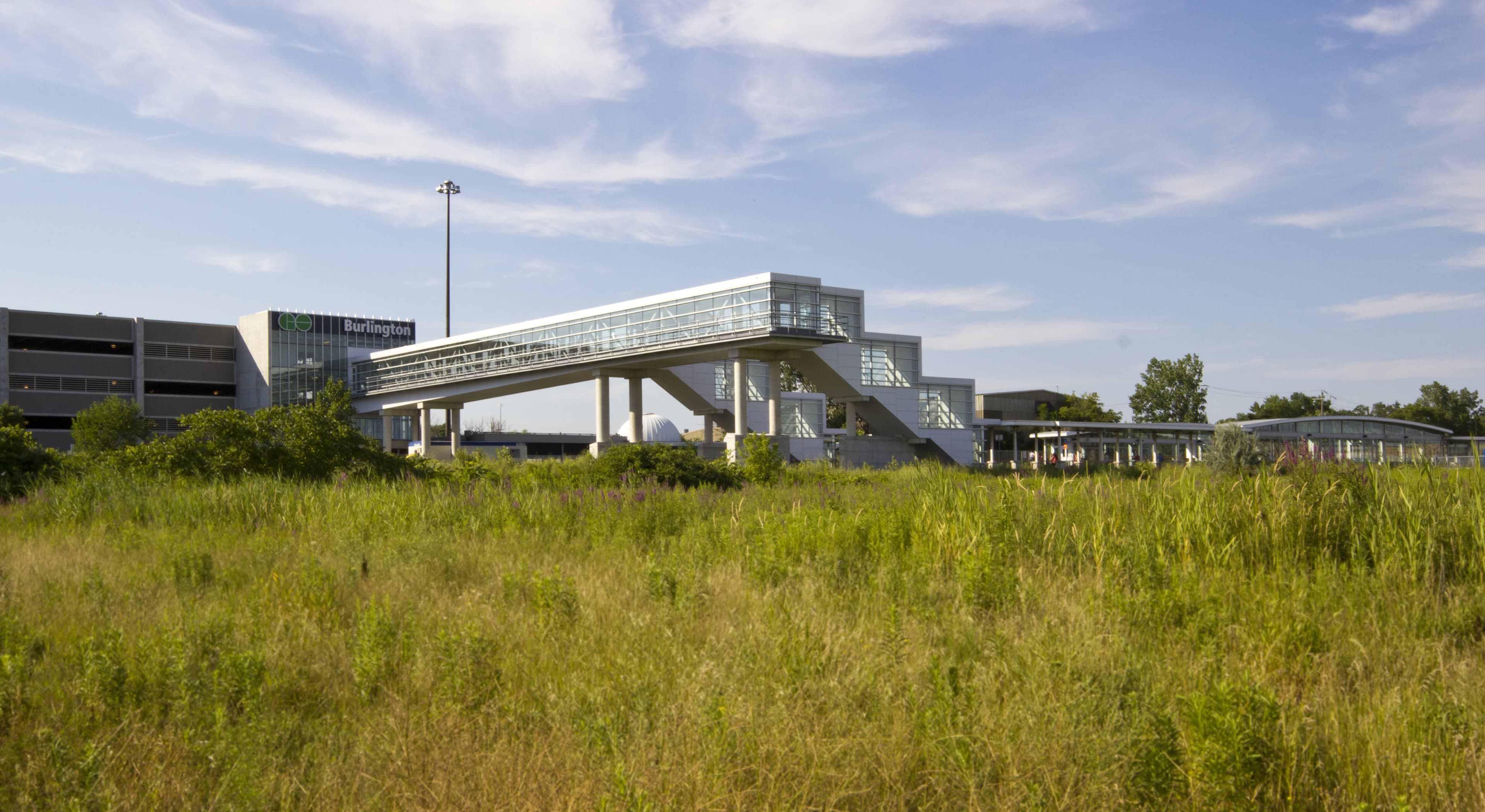 An enclosed pedestrian bridge and new stair towers connect the existing parking garage to the train platforms, spanning the CN main line. The structure was shop-fabricated in sections, assembled on site, and lifted into place overnight onto concrete piers. Its sloping floor provides clearance for trains, while a cantilevered catwalk accommodates maintenance. Clad in white aluminum panels with curtainwall, the bridge aligns visually with the surrounding station buildings from the 1960s and 70s.