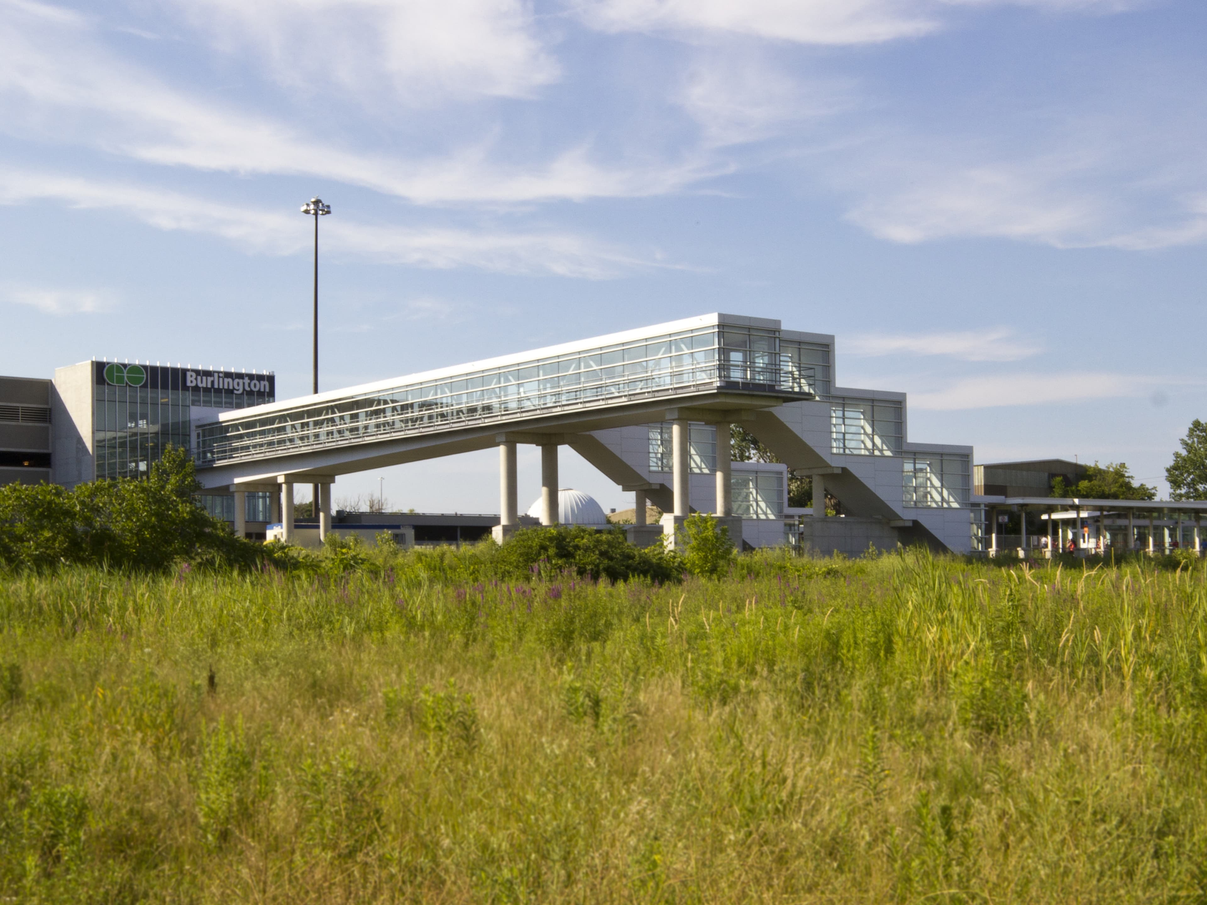An enclosed pedestrian bridge and new stair towers connect the existing parking garage to the train platforms, spanning the CN main line. The structure was shop-fabricated in sections, assembled on site, and lifted into place overnight onto concrete piers. Its sloping floor provides clearance for trains, while a cantilevered catwalk accommodates maintenance. Clad in white aluminum panels with curtainwall, the bridge aligns visually with the surrounding station buildings from the 1960s and 70s.