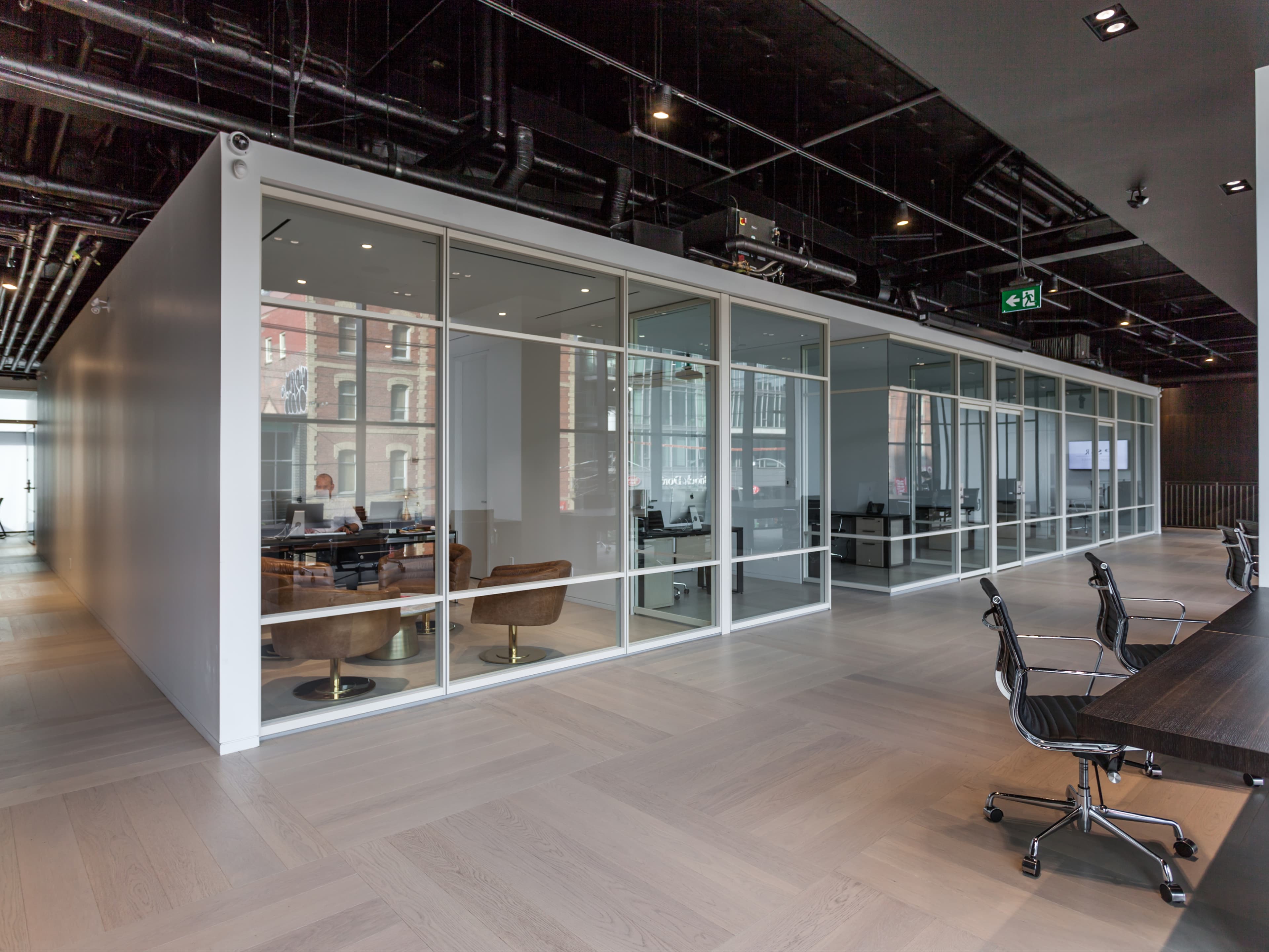This office for a Toronto real estate brokerage is organized across two levels, with a ground floor lobby and work areas located above. A lit stair leads to a reception hub, with open workstations along the glazed facade and a suite of glass-fronted meeting rooms and a boardroom arranged as freestanding volumes. Pale oak floors, white-framed partitions, dark metal detailing, and a matte black ceiling with warm integrated lighting create a calm, contemporary environment suited to client-facing work.